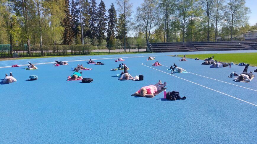 Urheilukentällä auringon paisteessa ryhmäliikuntatuntilaisia rentoutumassa. Kentän pinta on sininen, taivas on sininen, kirkas ja aurinkoinen.