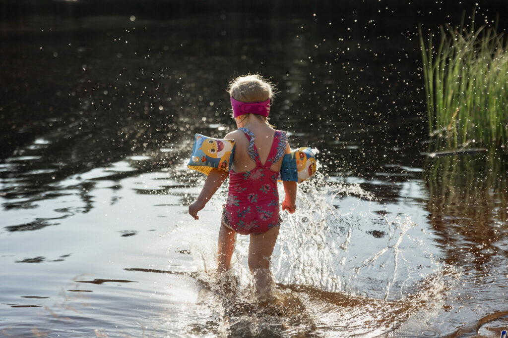 Pieni tyttö punaisessa uimapuvussa ja uimakellukkeet käsivarsissa kahlaa rannalla.