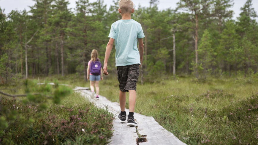 Tyttö ja poika kävelevät peräkkäin pitkospuilla. Taustalla on metsäinen maisema. Tytöllä on reppu selässä ja hän menee etummaisena. Poika kävelee perässä.
