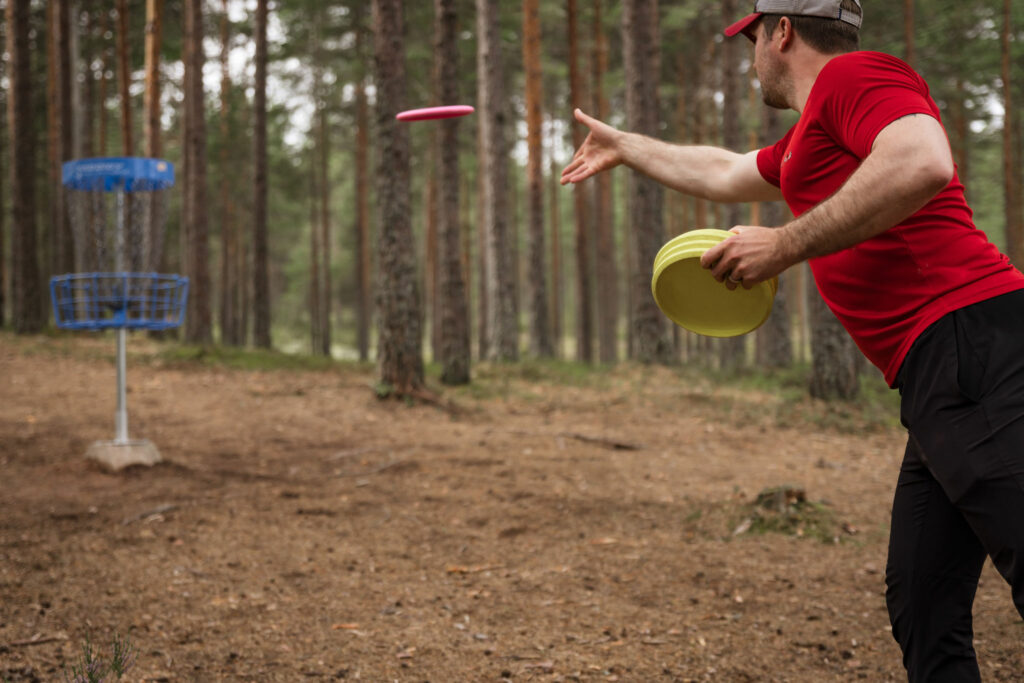 Frisbeegolffari heittää frisbeetä koriin metsäisessa maastossa.